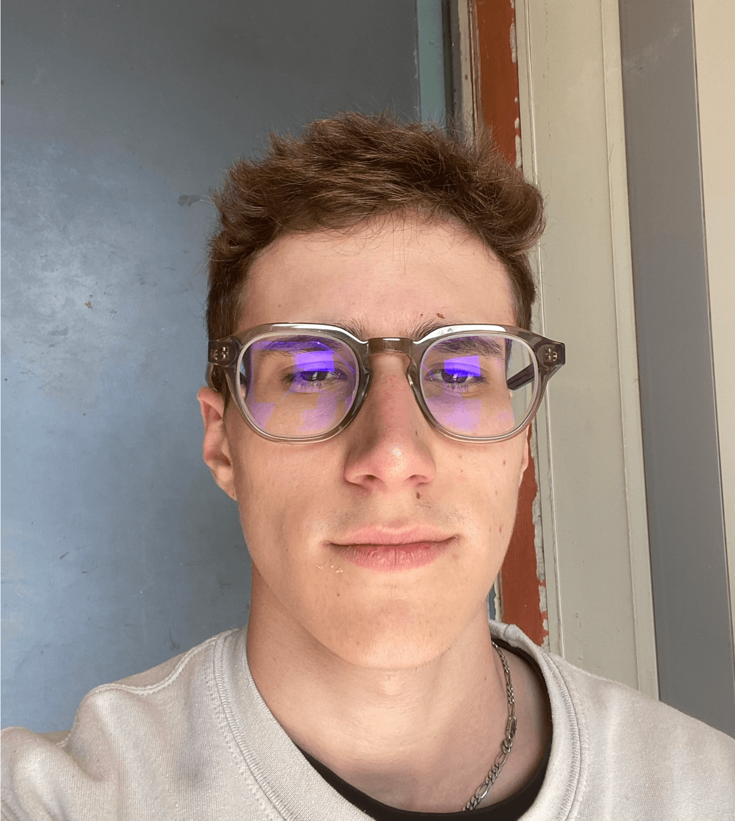 Young man with messy brown hair wearing thick clear glasses and a light-colored sweatshirt.